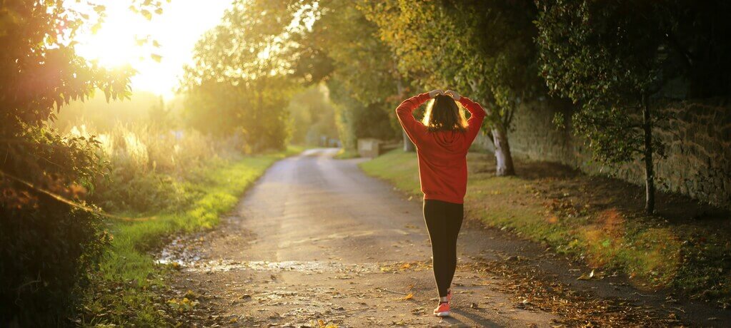 Une femme en tenue de sport marchant sur un chemin bordé d’arbres, baignée par la lumière du soleil. L’image symbolise l’écoute des sensations corporelles et la connexion avec soi-même dans un environnement naturel.